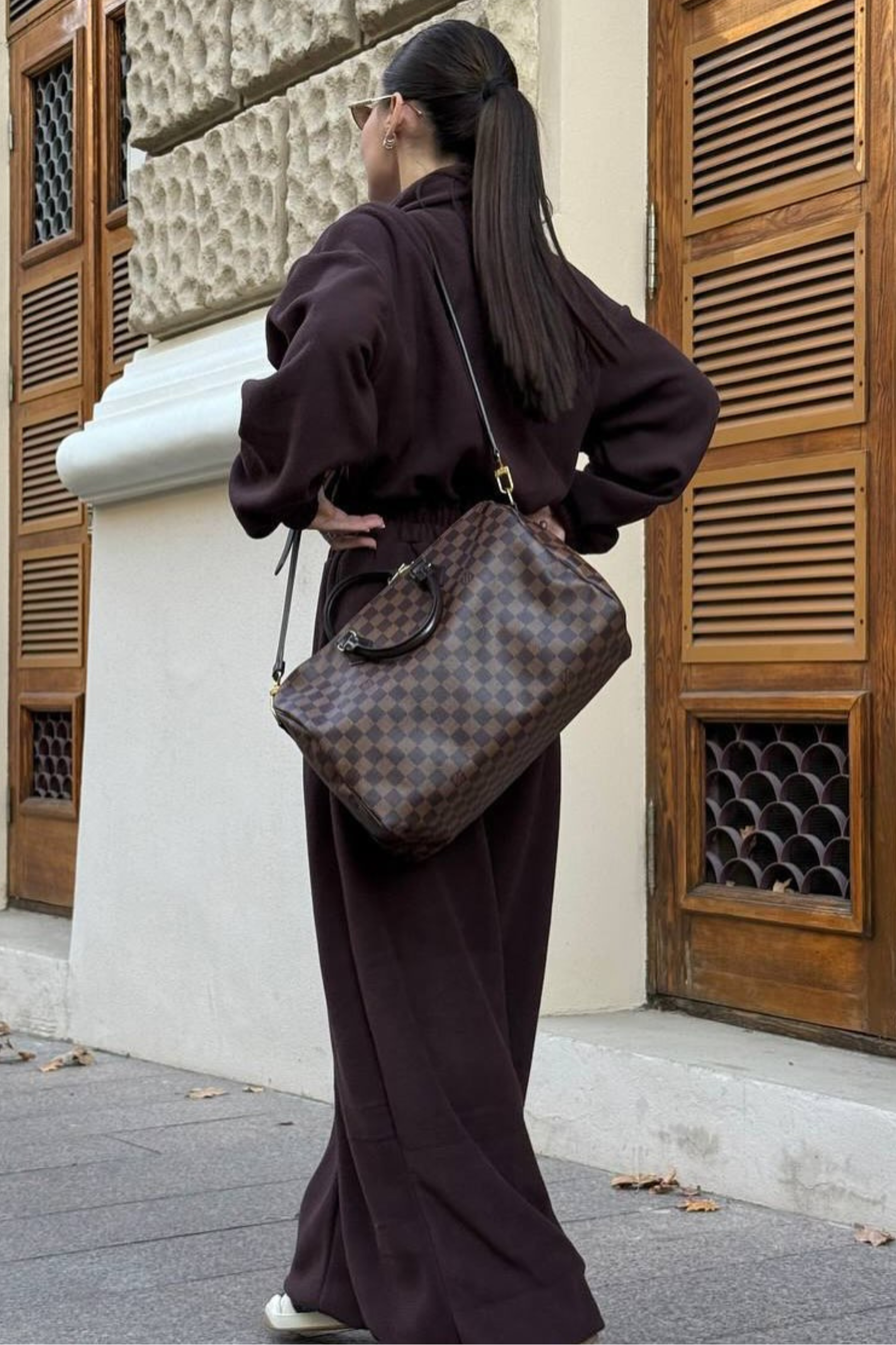 Person in a dark outfit with a patterned handbag standing against a building with wooden shutters.